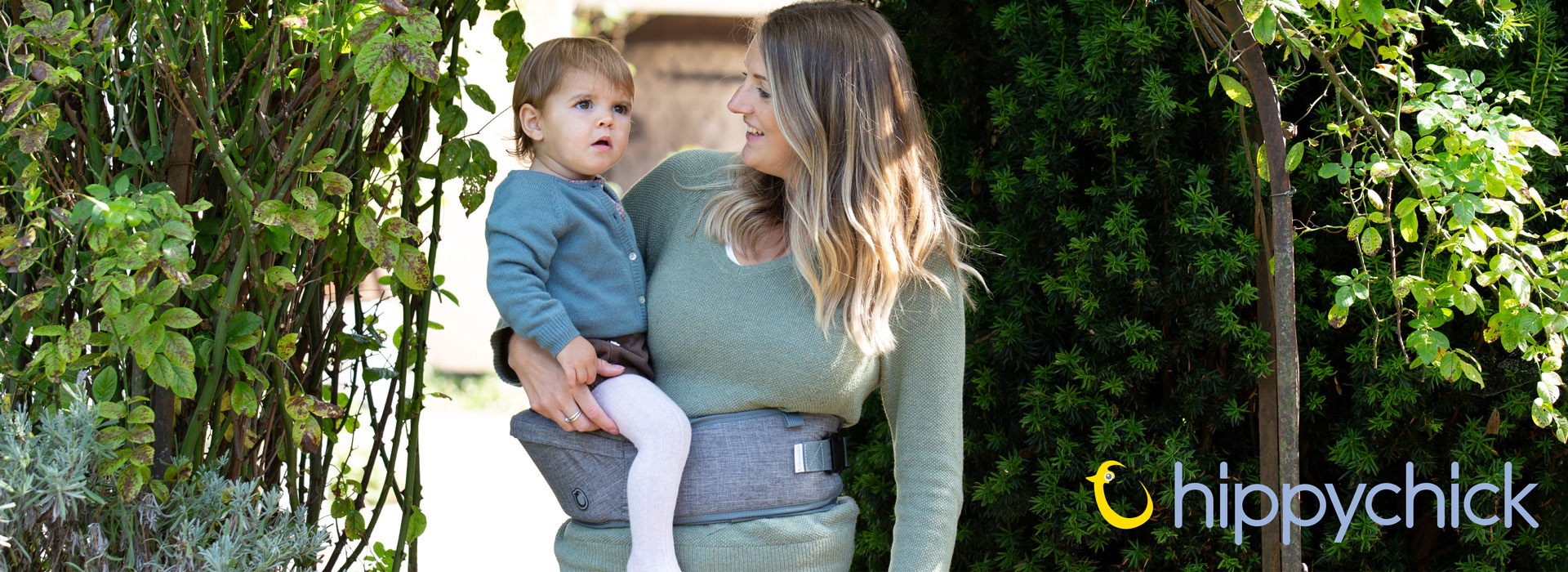 Kvinde bærer et barn i bæresele i en have omgivet af grønne planter.