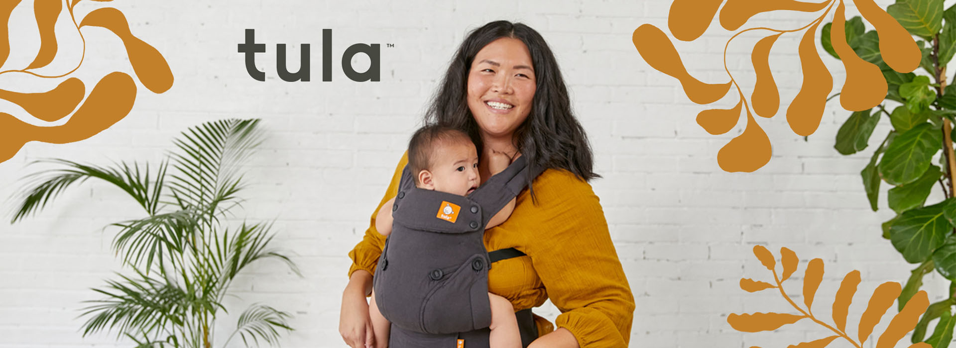 Kvinde bærer barn i grå bæreslynge mod hvid væg med Tula-logo.