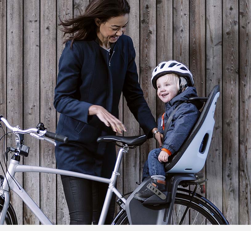 Kvinde med cykel, barn i cykelsæde har hjelm og smiler.