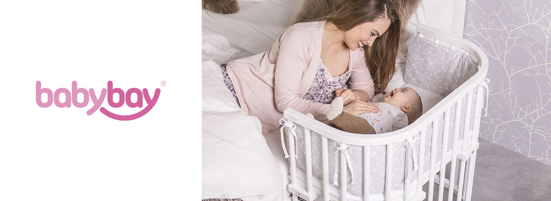 Mor smiler til baby i grå/hvid tremmeseng ved sengekant.