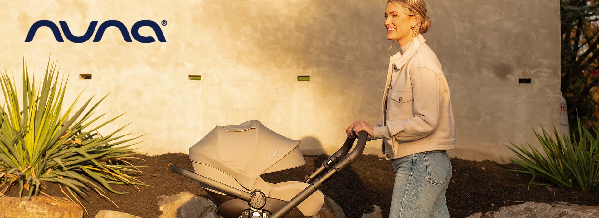 Kvinde i jakke går med barnevogn i solskin.