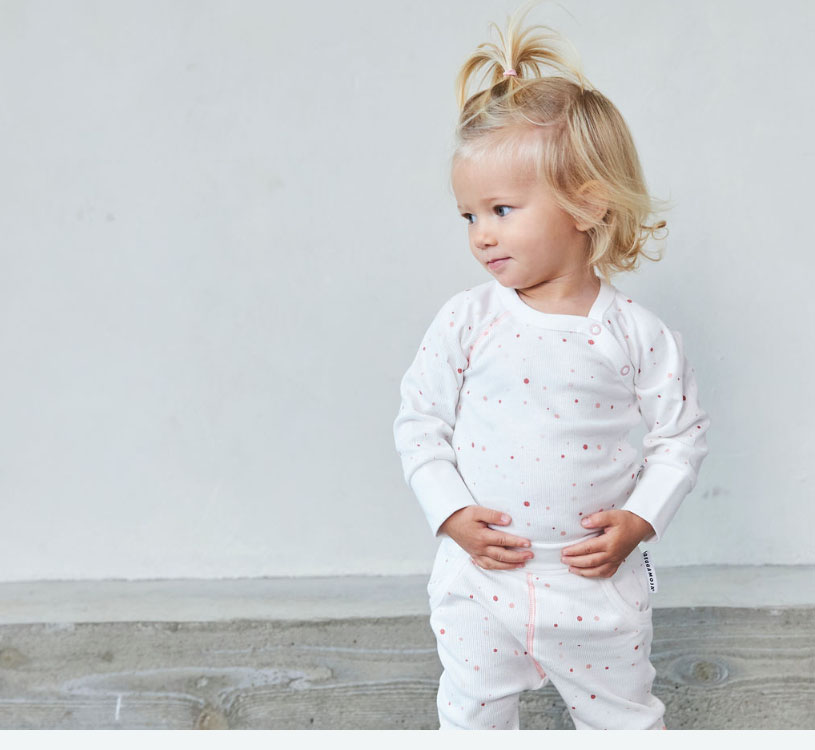Lille barn i hvid pyjamas med prikker, står mod en grå baggrund.