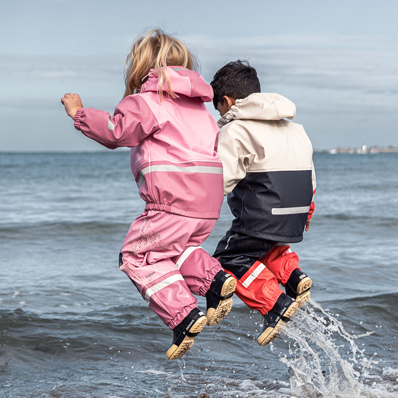 Barn i gul regnhat og grøn regntøj går på stenstrand.