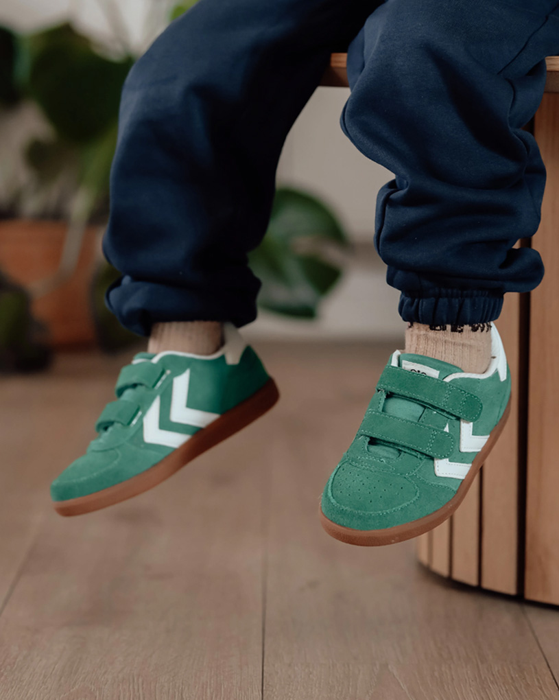 Barn i grønne sko med velcro og hvide detaljer på en træbænk.