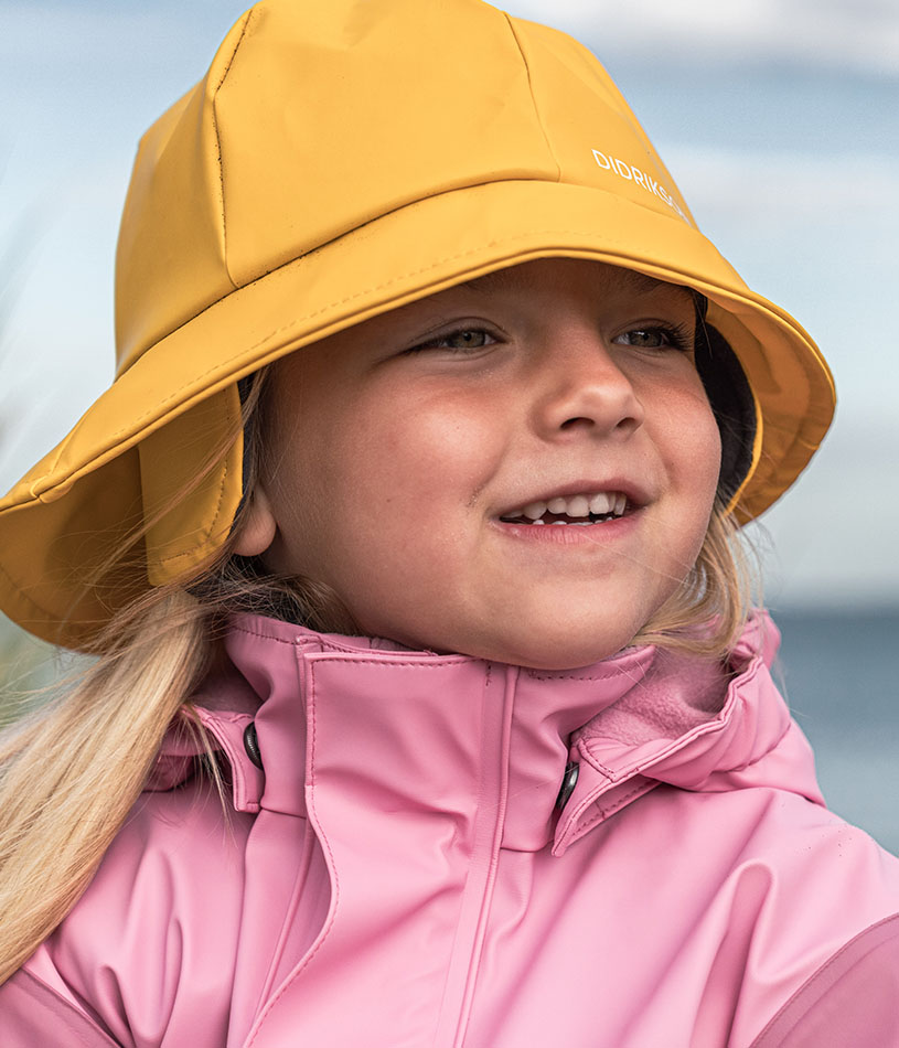 Barn i pink regnfrakke og hat smiler udendørs.