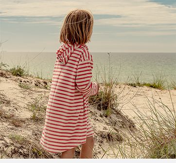 Barn i rød- og hvidstribet trøje står på stranden og kigger mod havet.