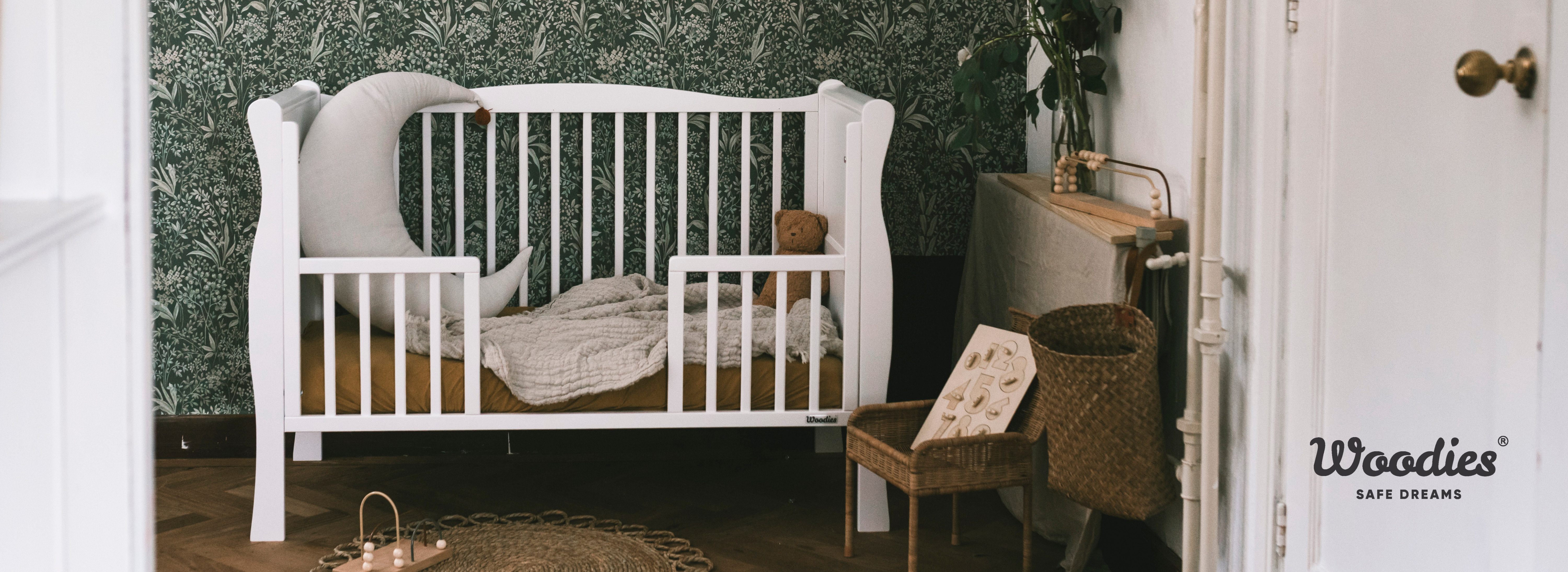 Hvid tremmeseng med bløde puder foran blomstret tapet i børneværelse.