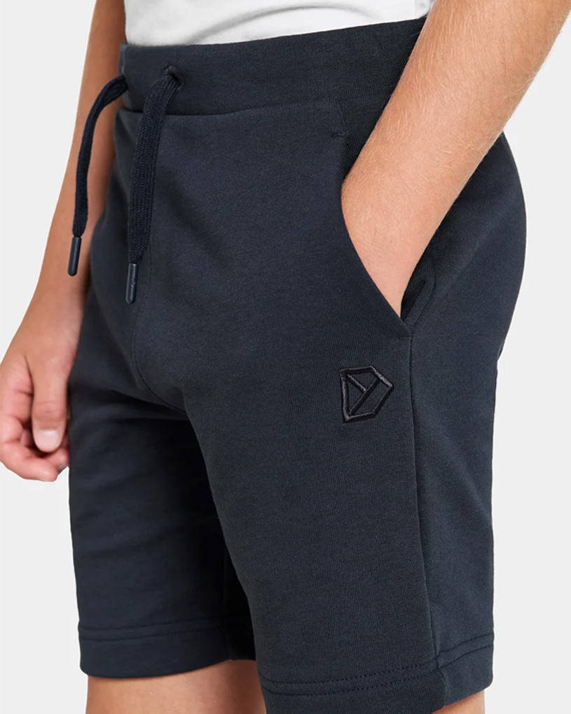 Nærbillede af en person iført mørkeblå shorts med lommer og et logo på venstre ben.