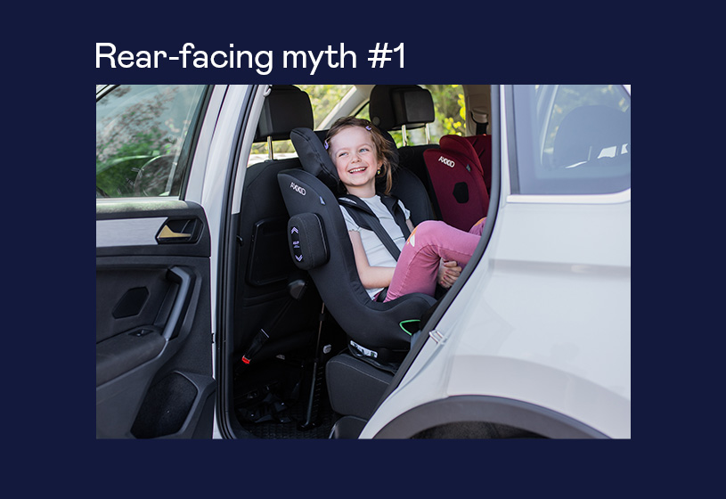 Barn i autostol baglæns i bilens bagsæde, smilende.