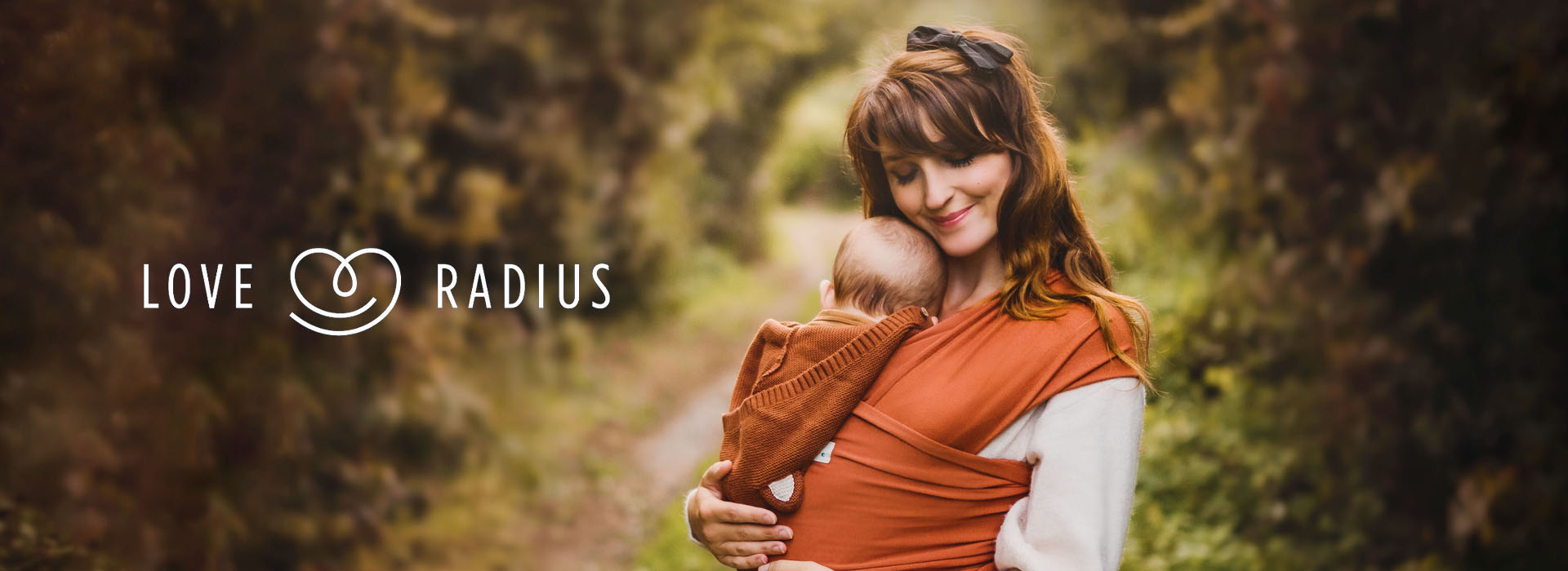 Kvinde holder baby i sjal i skovområde, logo til venstre.