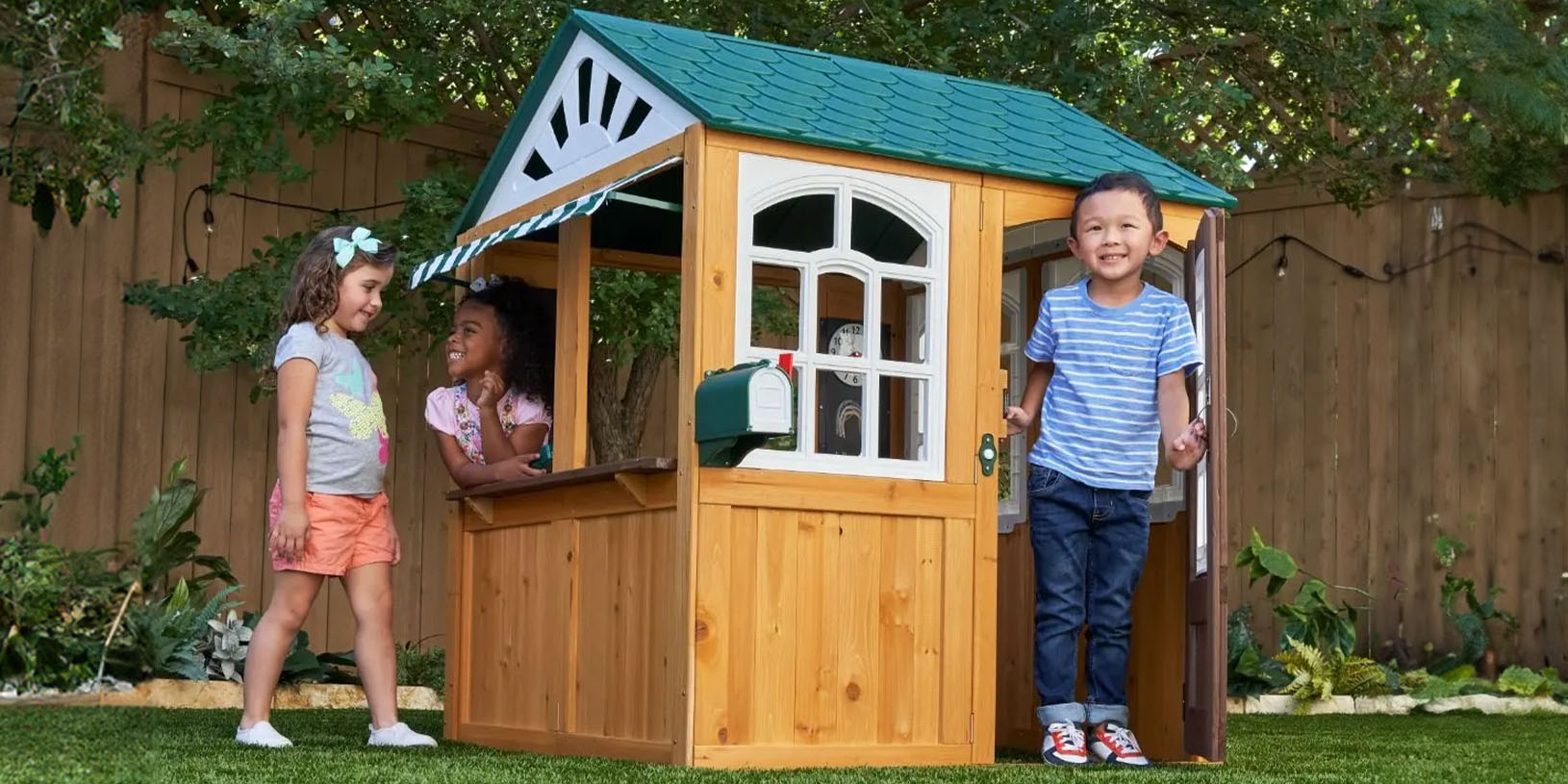 Tre børn leger ved et lille legehus af træ i en have.