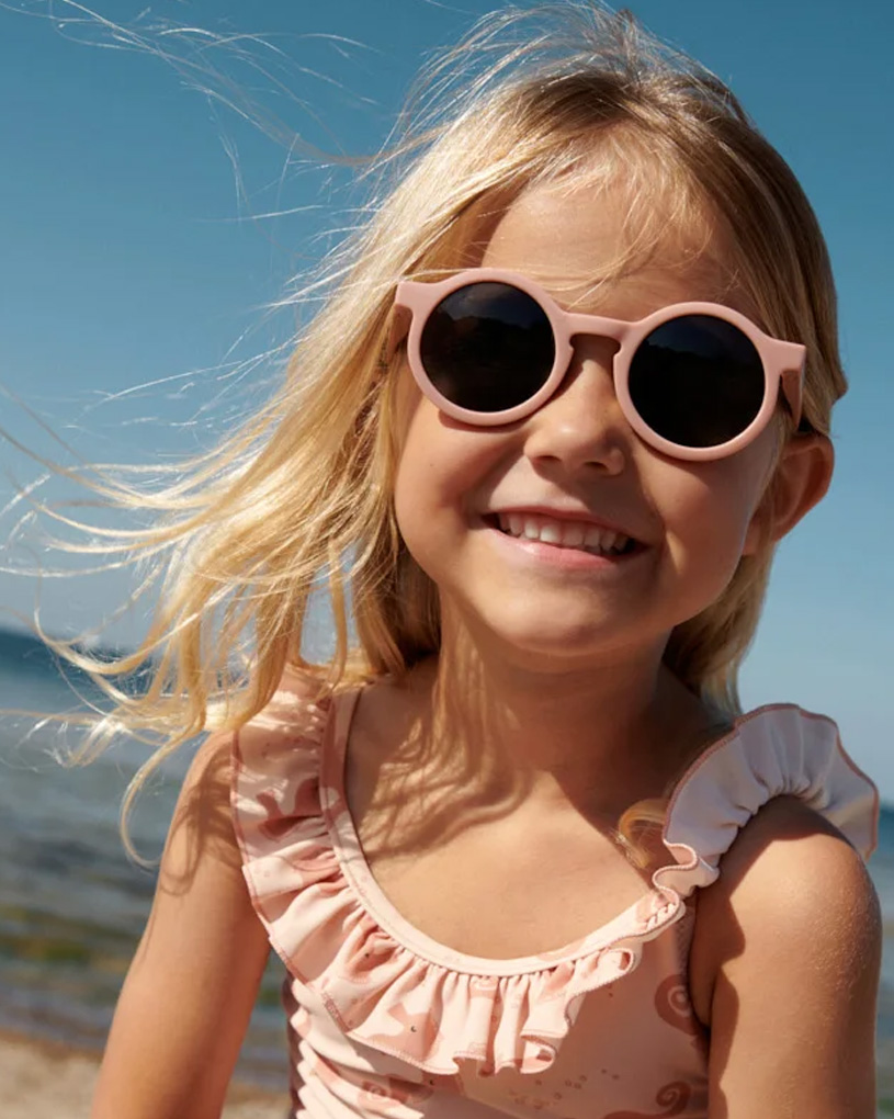 Smilende barn med solbriller på stranden under blå himmel.