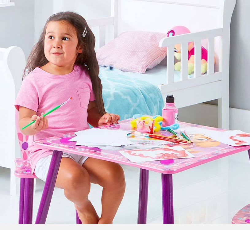 Pige i pink trøje maler ved lyserødt bord med pensel.