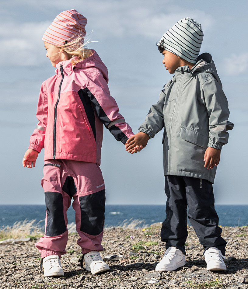 Barn i lyserød regntøj med en skovl og spand ved vandet.