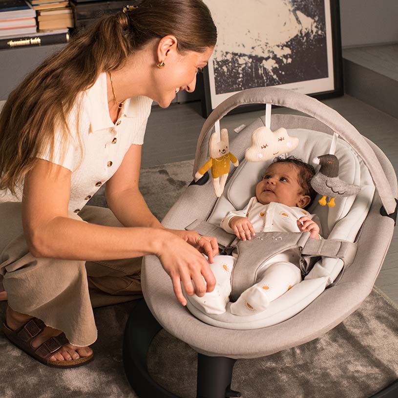 Kvinde smiler til baby i skråstol med legetøj.