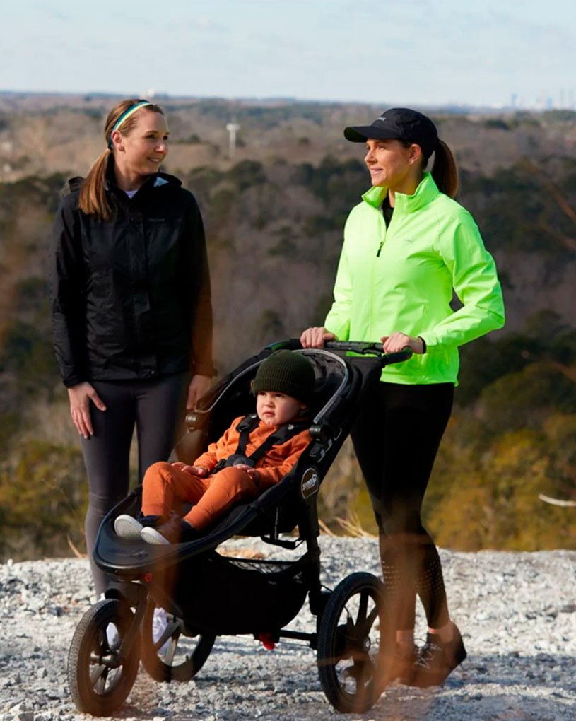 To kvinder med en barnevogn på en grusvej med udsigt over en skov.