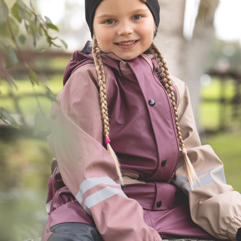Smilende barn i farverig regnjakke, sidder ude i grønt område.