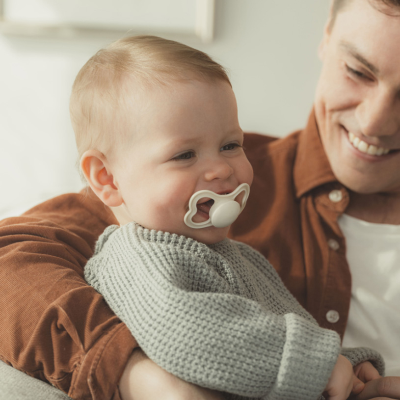 Baby med sut i armene på en smilende voksen.