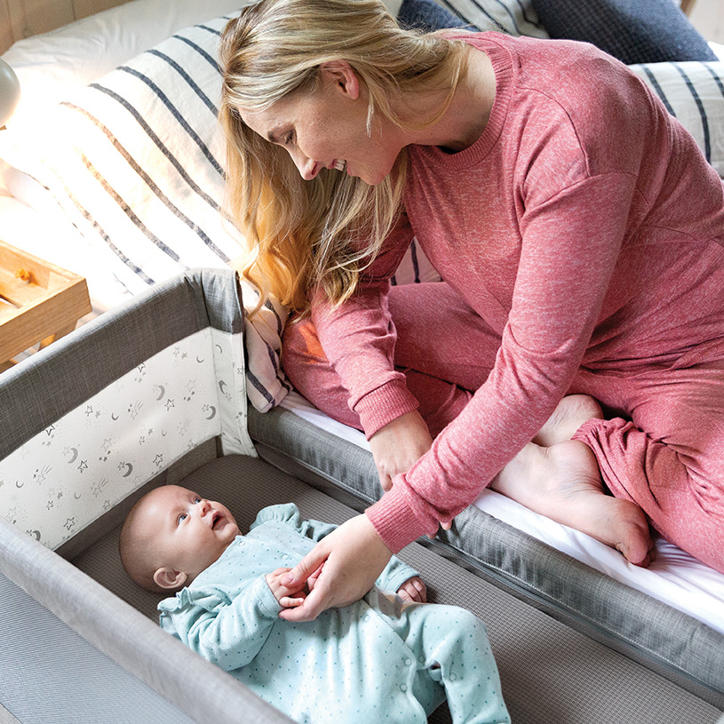 Baby i gyngestol med legetøj, pige sidder ved bord i baggrunden.