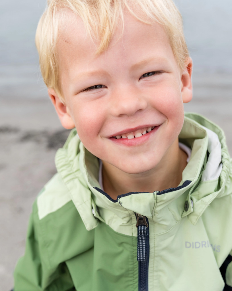 Smilende barn i grøn jakke ved strand.