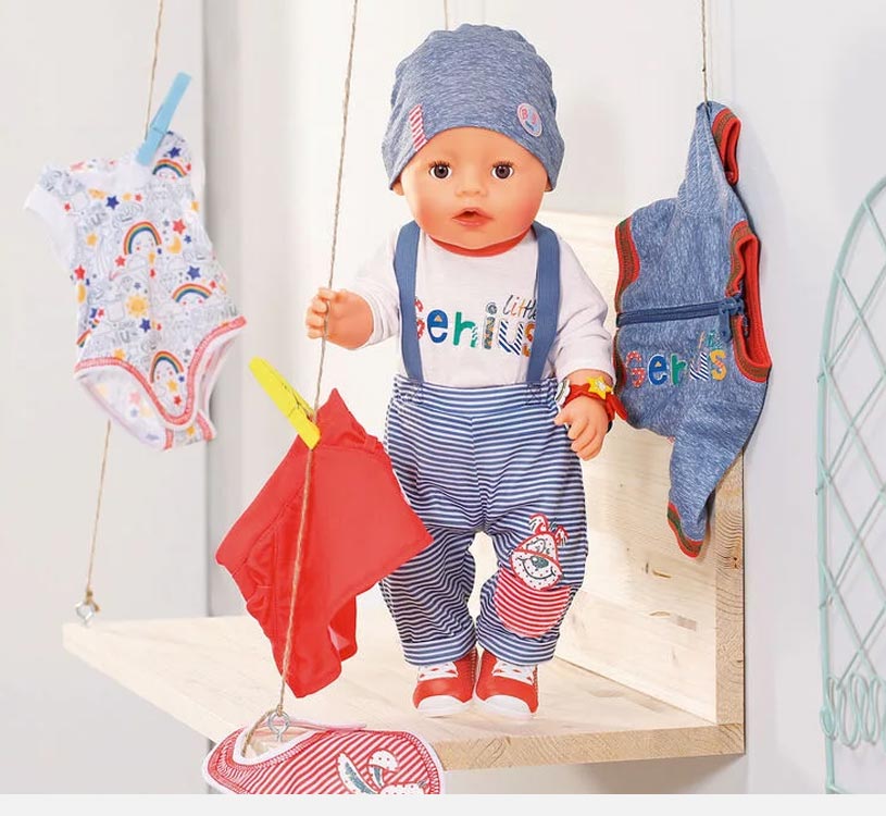 Dukke med stribede bukser og hue på hylde, omgivet af babyklæder.