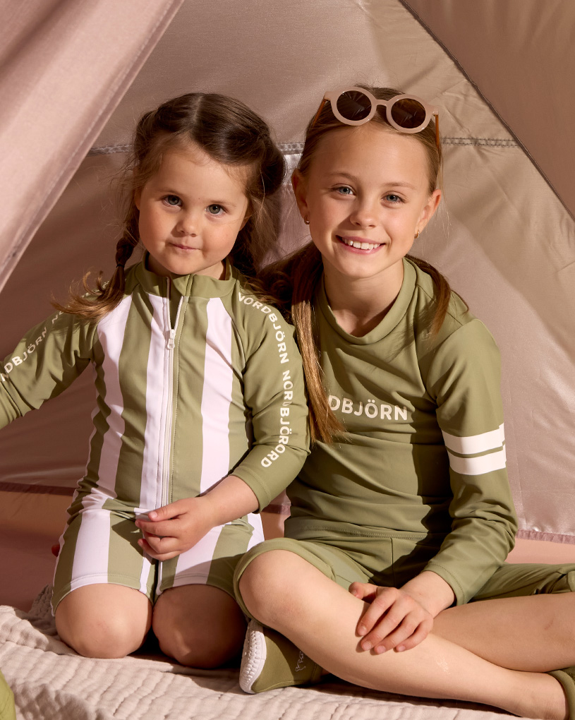 To børn i grønne UV-dragter smiler inde i et telt.