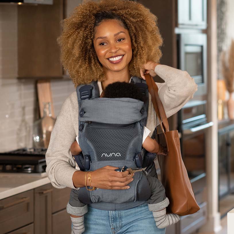 Kvinde bærer et barn i en sele og holder en brun taske.