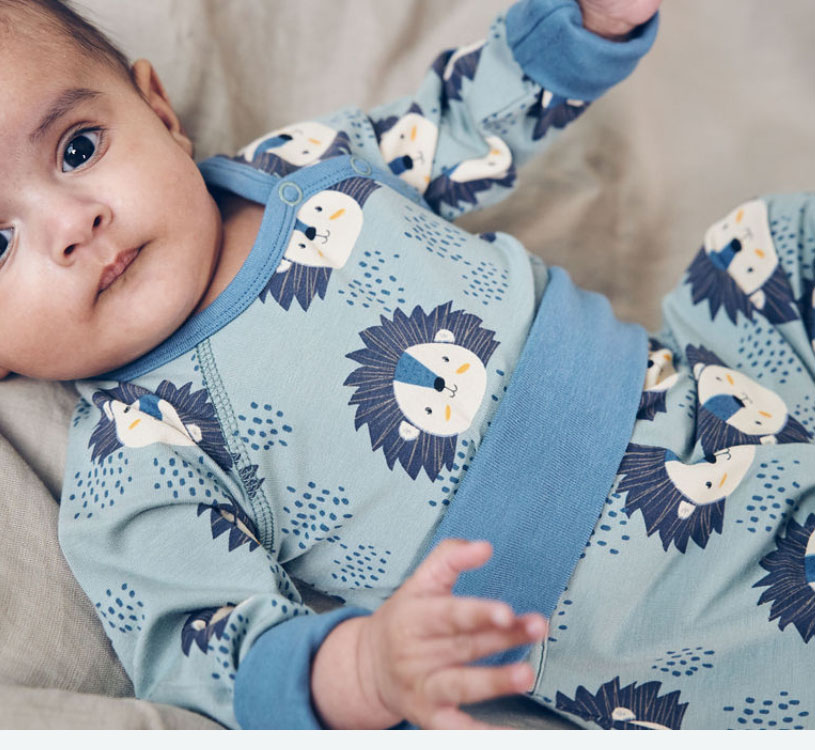 Baby i blå pyjamas med løvemønster, liggende på beige pude.